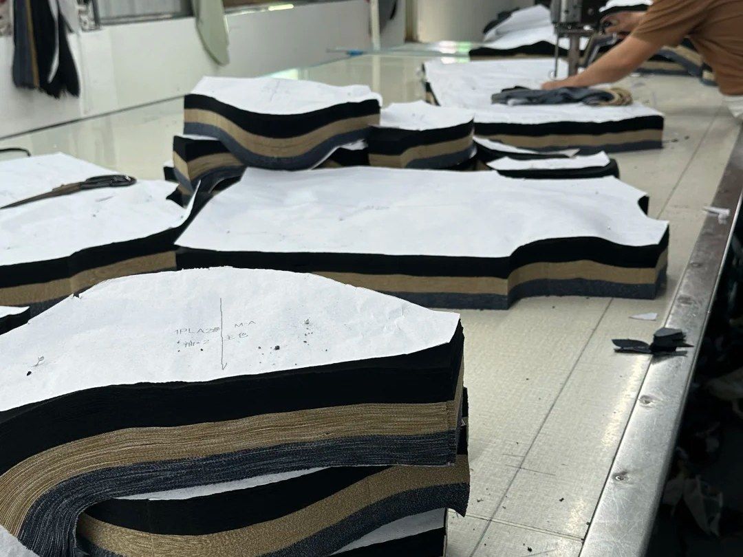 Stacks of cut garment panels topped with paper markers on our cutting table, a cutter working in the background