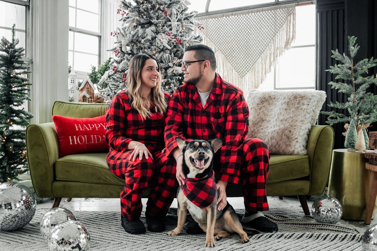 Couple in matching red buffalo-check pajama sets sitting with a dog in a coordinating bandana
