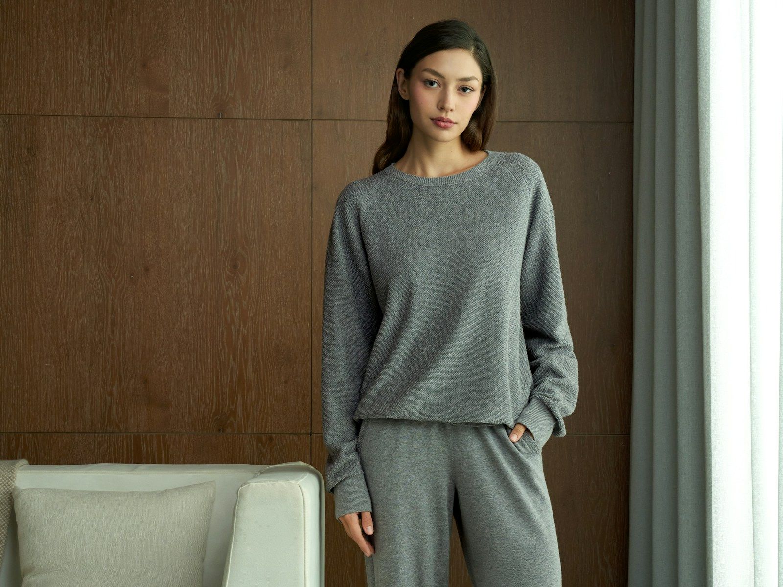 Woman standing by a window in a matched grey crew-neck knit lounge top and pants, one hand in the side pocket
