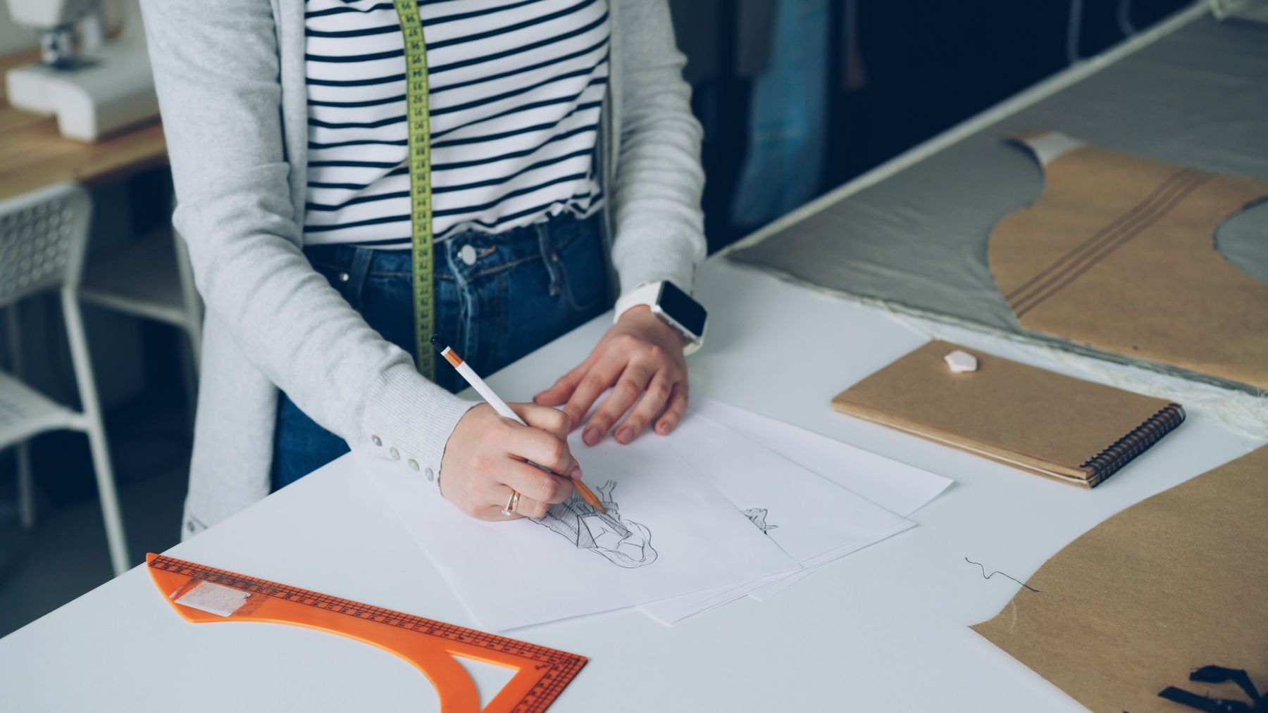 Designer sketching a garment at a desk with pattern paper, a ruler and a tape measure