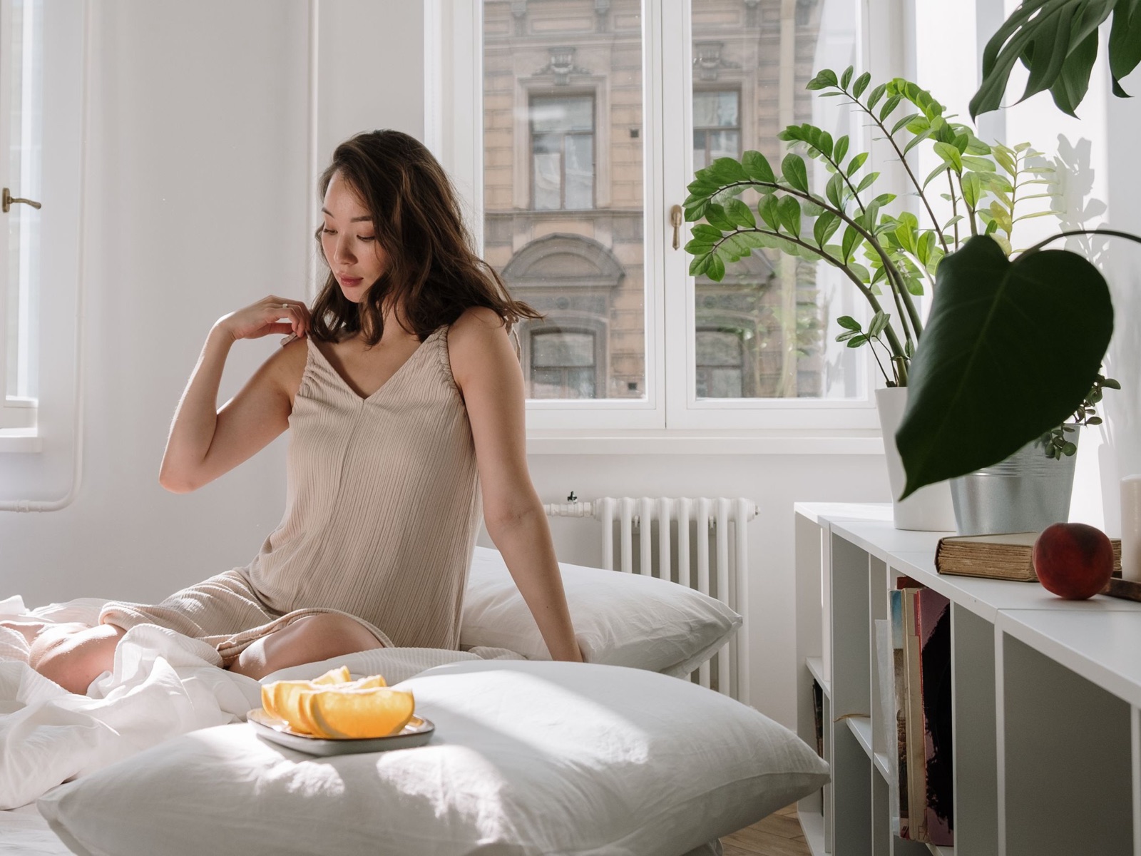 Woman in a beige ribbed nightdress sitting on a bed in a sunlit bedroom